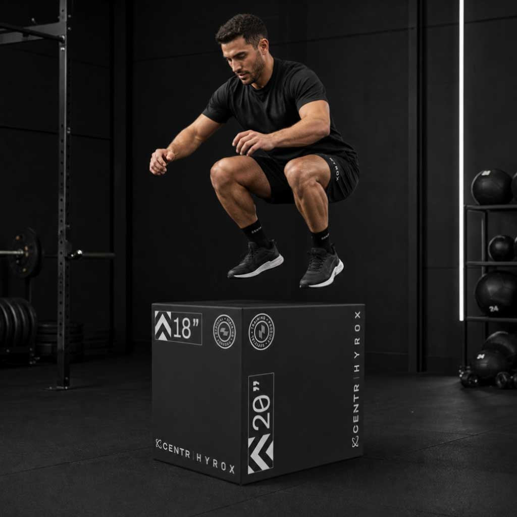 Man in black sportswear jumps above a black foam plyometric box in a dark training space.