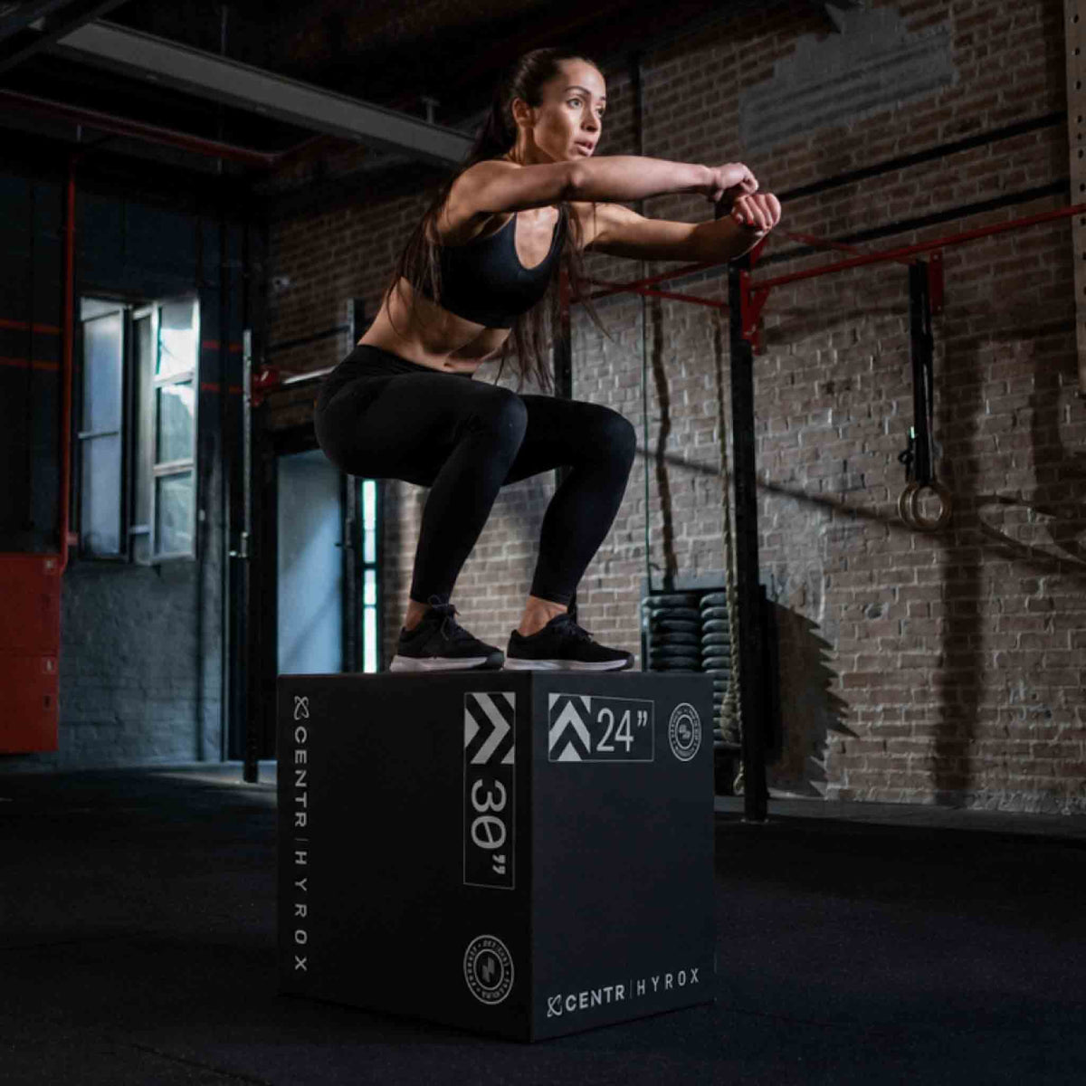 Woman in black sportswear jumps onto a black foam plyometric box in a dark training space.