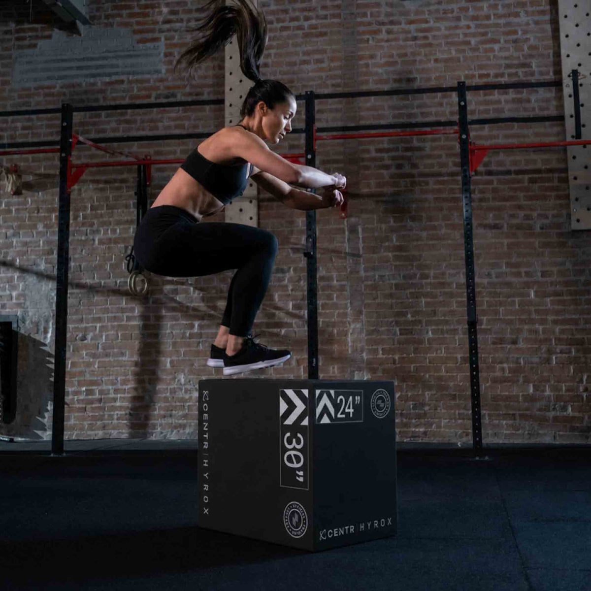 Woman in black sportswear jumps onto a black foam plyometric box in a training space with a brick wall.