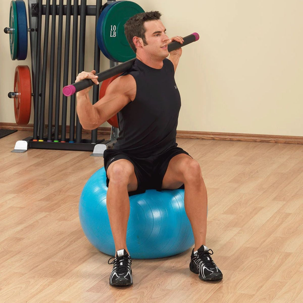 Man zit op een blauwe fitnessbal met een zwarte stang op zijn schouders; achter hem staan halterschijven en een rek.