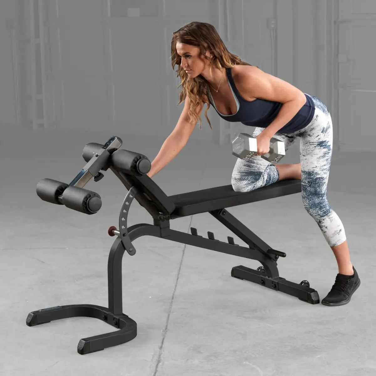 Vrouw steunt met één hand en knie op een trainingsbank en roeit met een halter in de andere hand.