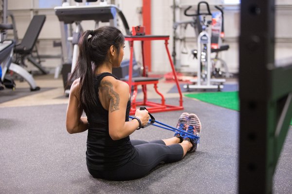 Vrouw zit op de fitnessvloer en trekt met beide handen een blauwe weerstandsband naar haar toe.