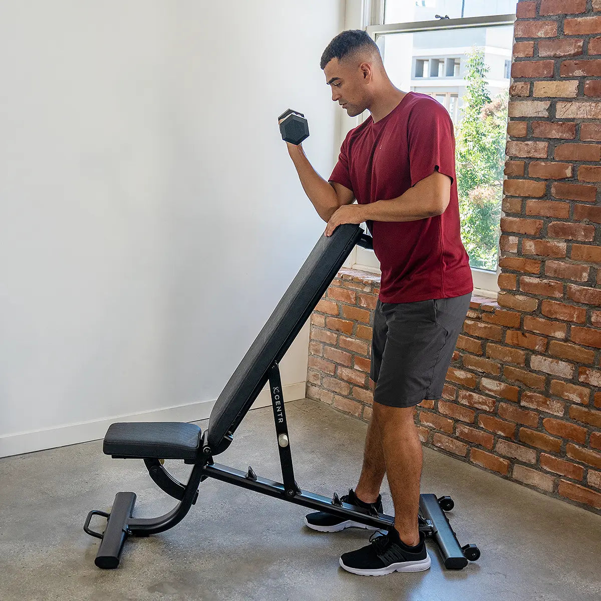 Man in rood shirt staat naast een schuine trainingsbank en steunt met één hand op de rugleuning.