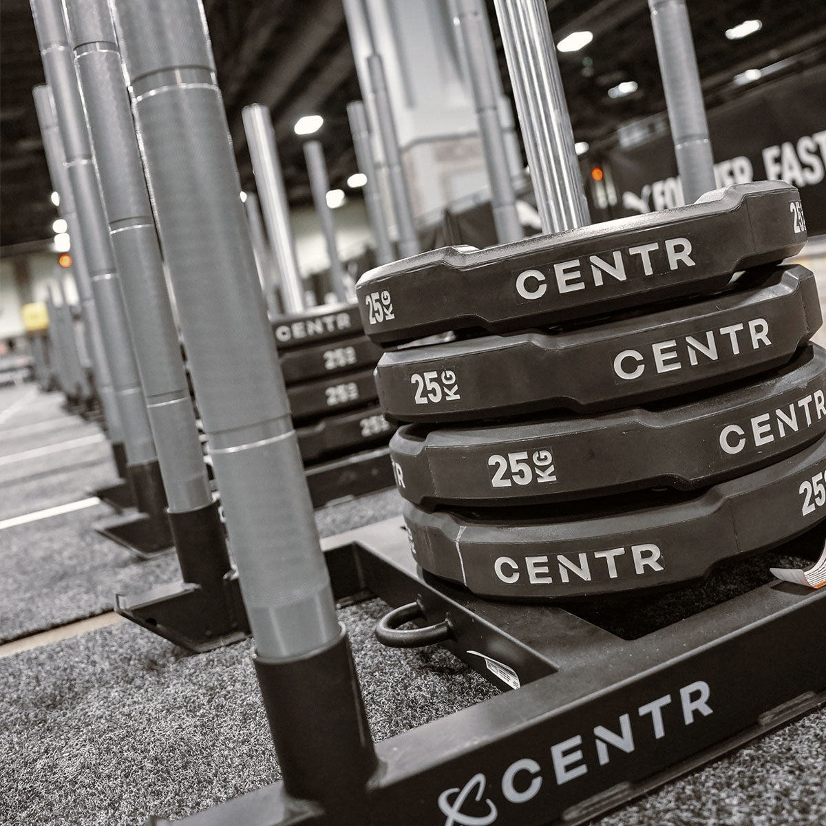 Stapel zwarte CENTR-bumperplates op houders in een fitnessruimte; metalen staanders en het opschrift ‘25 KG’ zijn zichtbaar.