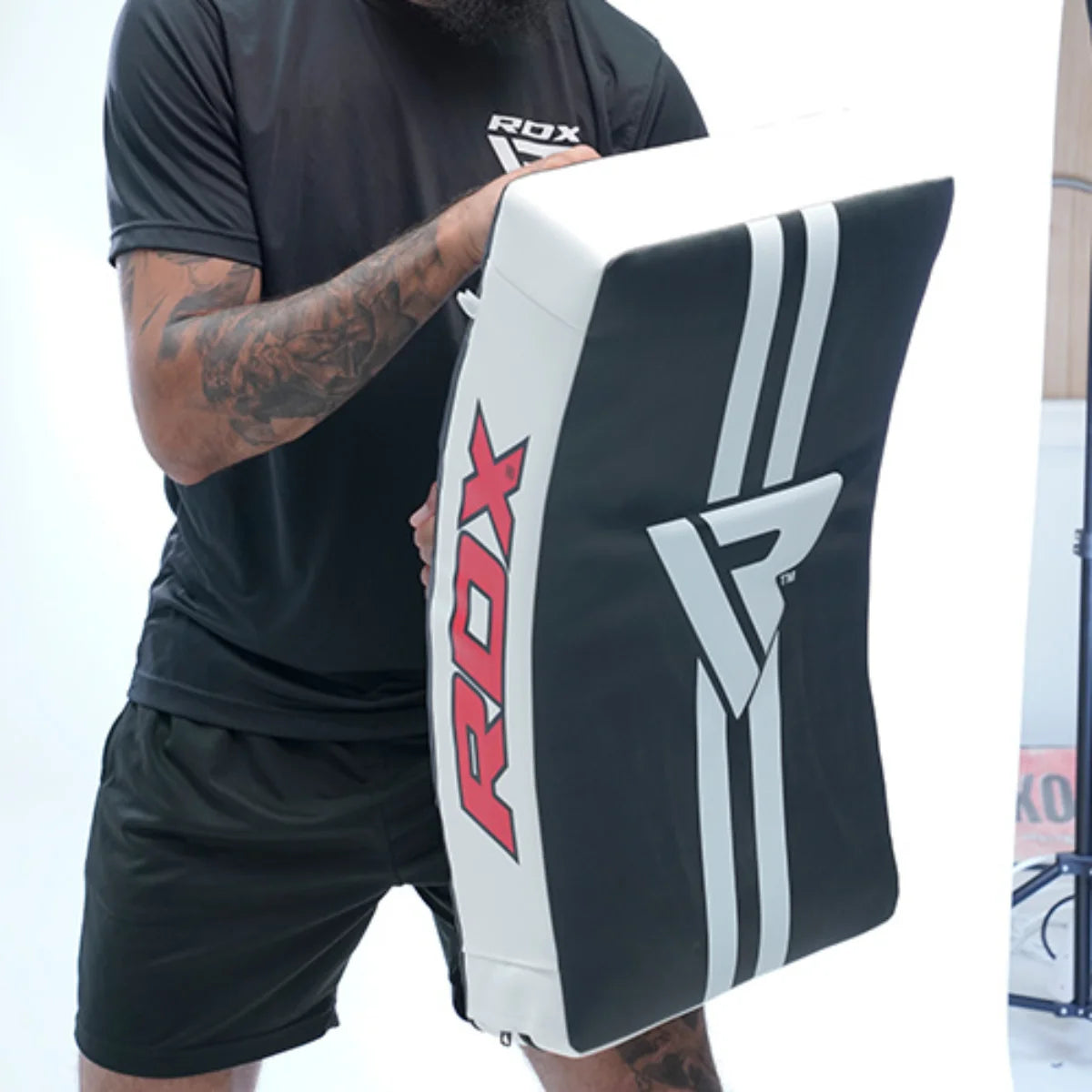 Persoon in zwarte sportkleding houdt een grote zwart-witte RDX-kickpad voor het lichaam; de onderarmen zijn zichtbaar getatoeëerd.