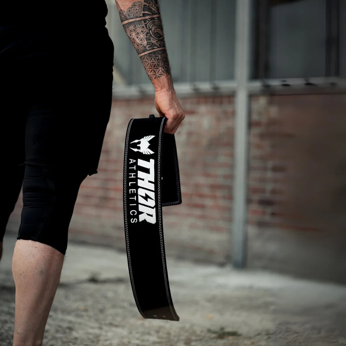 Hand houdt een zwarte riem met wit Thor Athletics-logo naast een sportbroek.