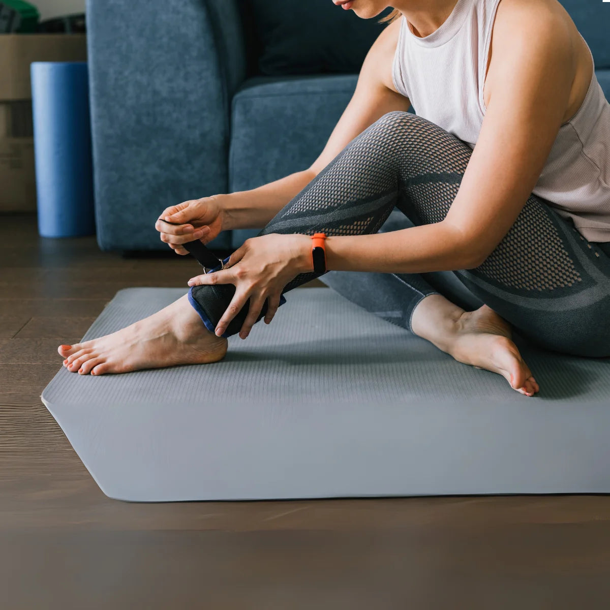 Een zittende vrouw maakt een zwart enkelgewicht vast op een fitnessmat; naast haar liggen een blauwe foamroller en een grijze bank.