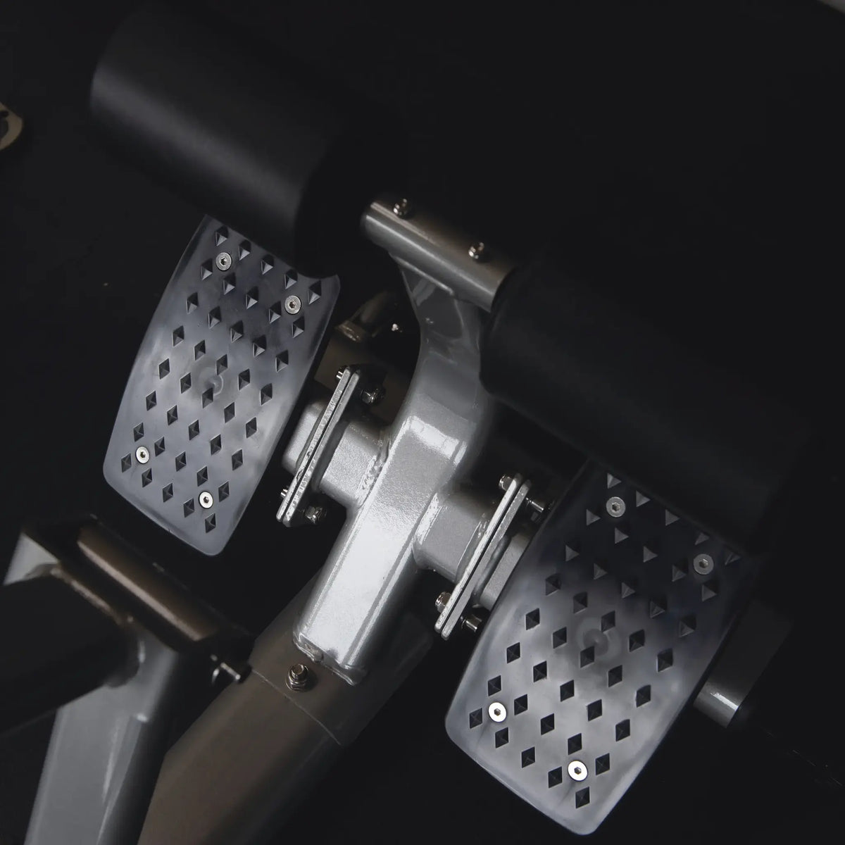 Close-up van twee metalen voetplaten met antislipgaten op een zwart fitnessapparaat.